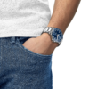A person wearing a silver watch with a blue dial, hand in pocket of blue jeans.