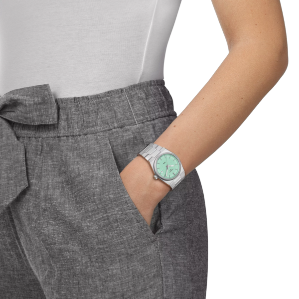 Woman wearing a mint green wristwatch with grey pants and white top.